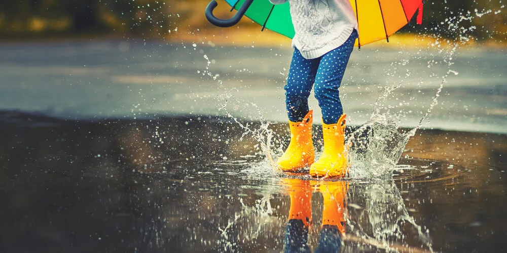 Kind mit gelben Gummistiefeln und buntem Regenschirm spring in eine Wasserpfütze