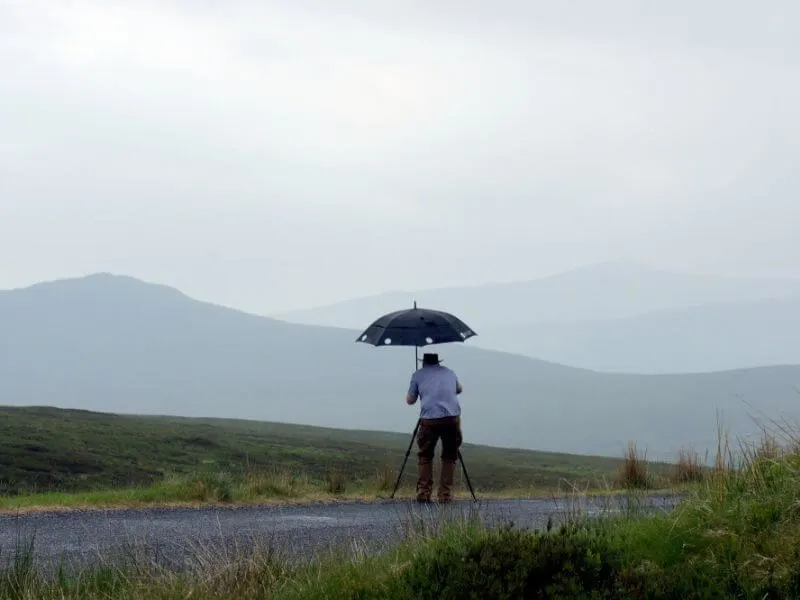 Man with tripod under an umbrella in the rain in Ireland.
